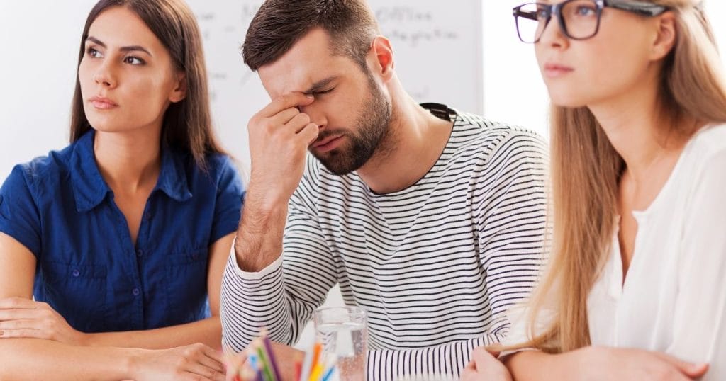 ragazzo fa fatica a concentrarsi , è seduto su una scrivania con due ragazze e si tiene la testa tra le mani